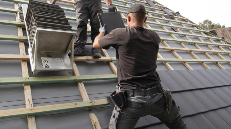 Zwei Handwerker verlegen Dachziegel auf einem geneigten Dach, während einer die Ziegel anreicht.