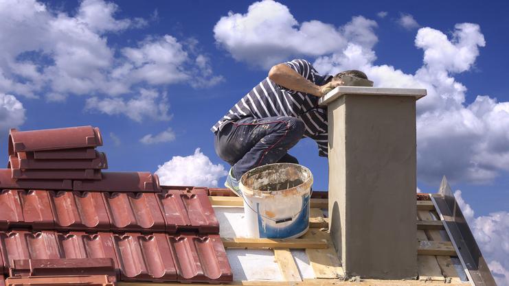 Handwerker in gestreiftem Shirt sitzt auf einem Dach und arbeitet am Mauerwerk eines Schornsteins unter wolkigem Himmel.