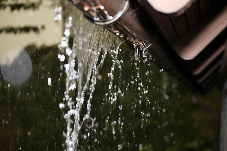 Wassertropfen fließen aus einem Regenblech herab, mit verschwommenem Hintergrund und sanfter Lichtreflexion.