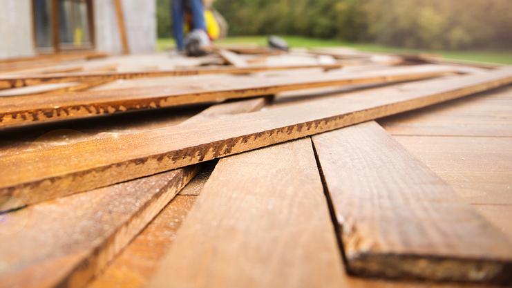 Holzdielen liegen auf einem Außenboden, während ein Arbeiter im Hintergrund mit einem Werkzeug hantiert.